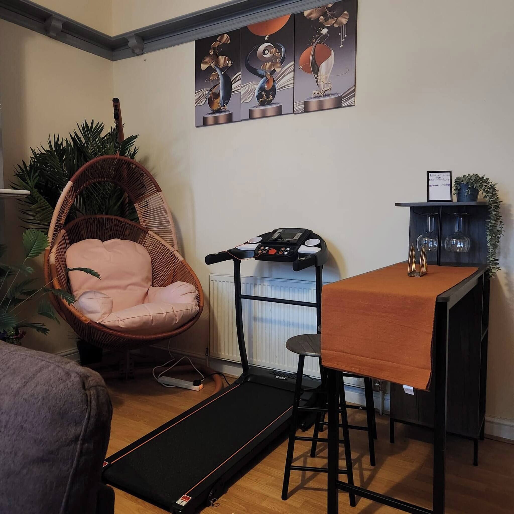 En kompakt opholdsstue med en polstret stol, et løbebånd, et lille bord med en bærbar computer og en radiator.