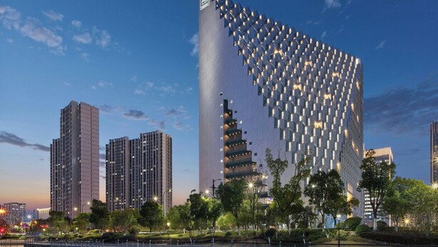 Un immeuble moderne de grande hauteur, doté d’une façade originale, entouré d’autres bâtiments imposants et d’un parc arboré.