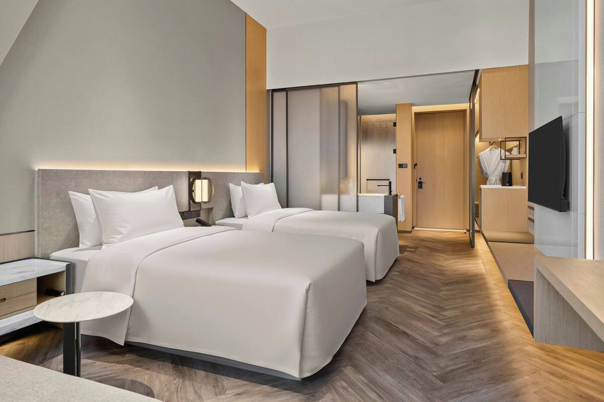 A modern hotel room with two beds, a small table, a TV, and a bathroom visible through a sliding door.