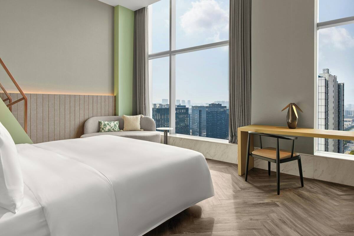 A modern hotel room with a large bed, a desk, and a view of the cityscape.