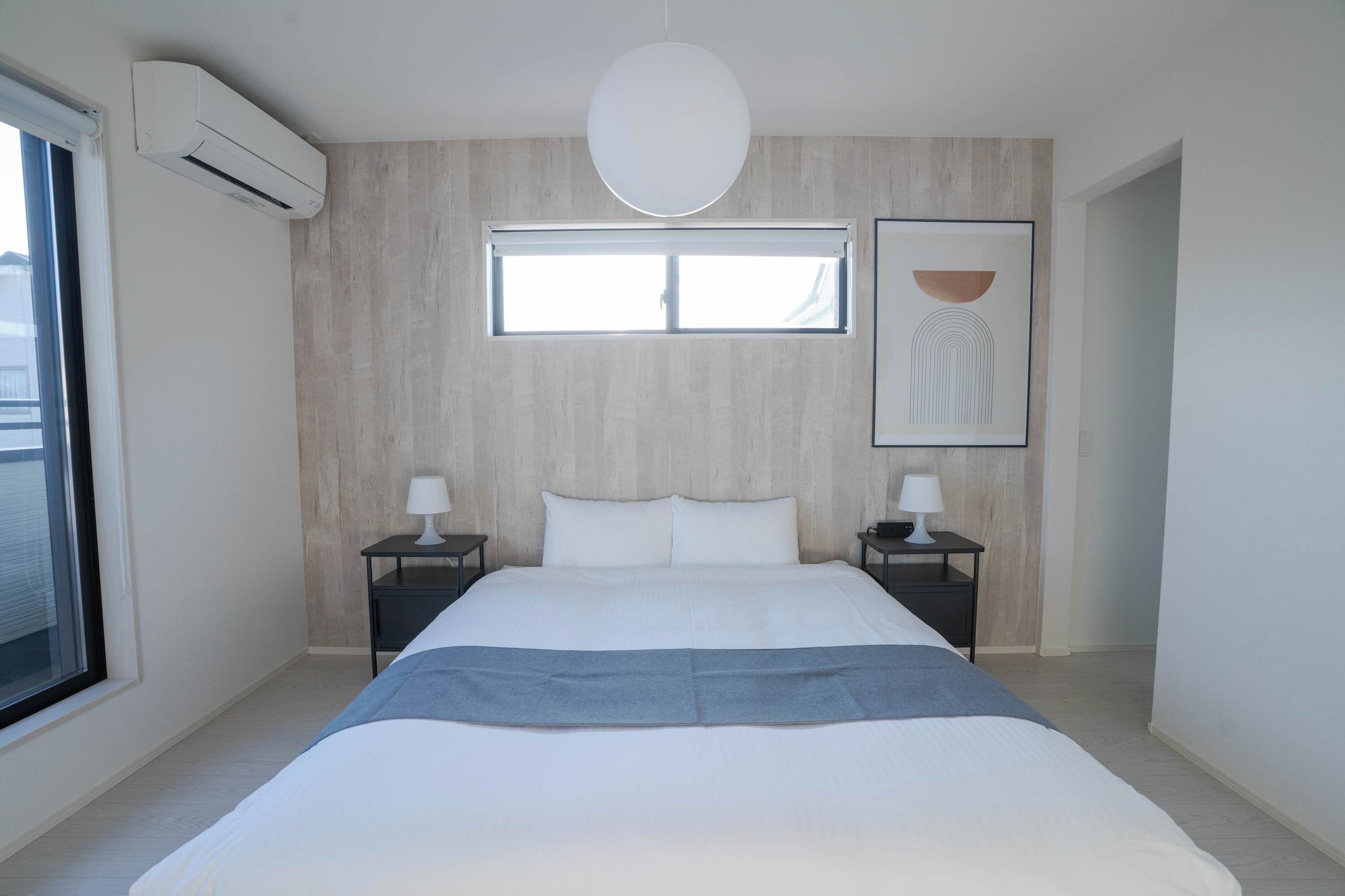 A modern bedroom with a large bed, two bedside tables with lamps, a window, and an air conditioning unit.