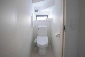 A small, white bathroom with a toilet, a window above the sink, and a light switch on the wall. - 4BR Modern Home Near Kumamoto Airport by Restavio (Ozu)