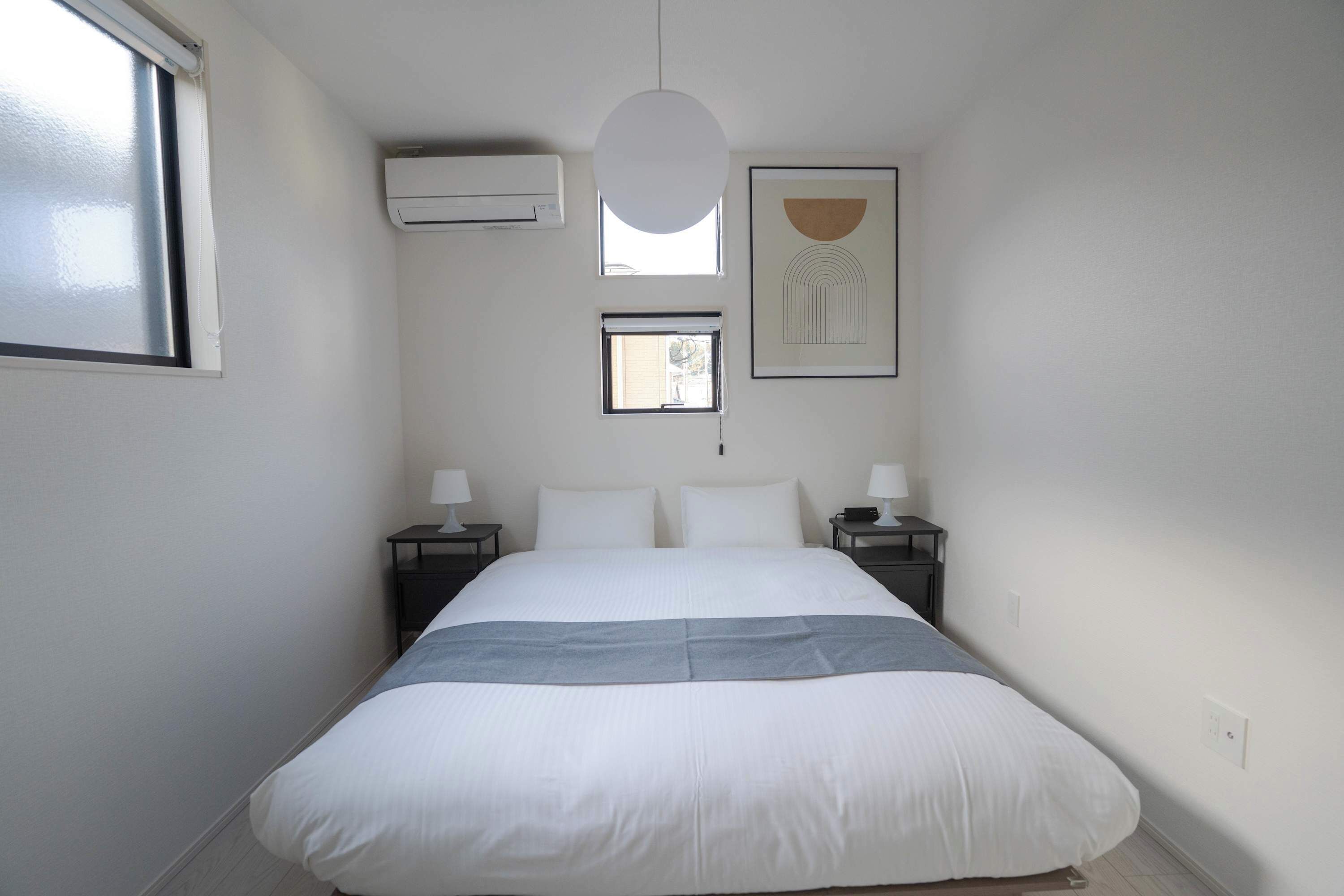 A bedroom with a large bed, two bedside tables, a hanging light, an air conditioner, and a window.