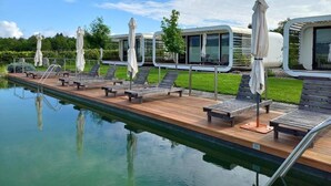 A poolside area with lounge chairs and umbrellas, a wooden deck, and a view of a modern building in the background. - Glamping Malerič with Natural Pool (Črnomelj)