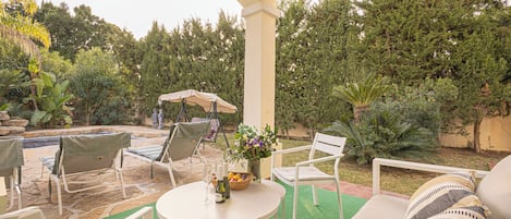 Une terrasse extérieure avec des meubles blancs, une table ronde sur laquelle sont disposées des boissons, et une piscine en arrière-plan.