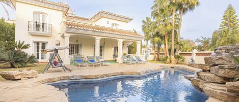 Una piscina con borde de baldosas, un patio con sillones de descanso y un edificio con techo y columnas de azulejos.