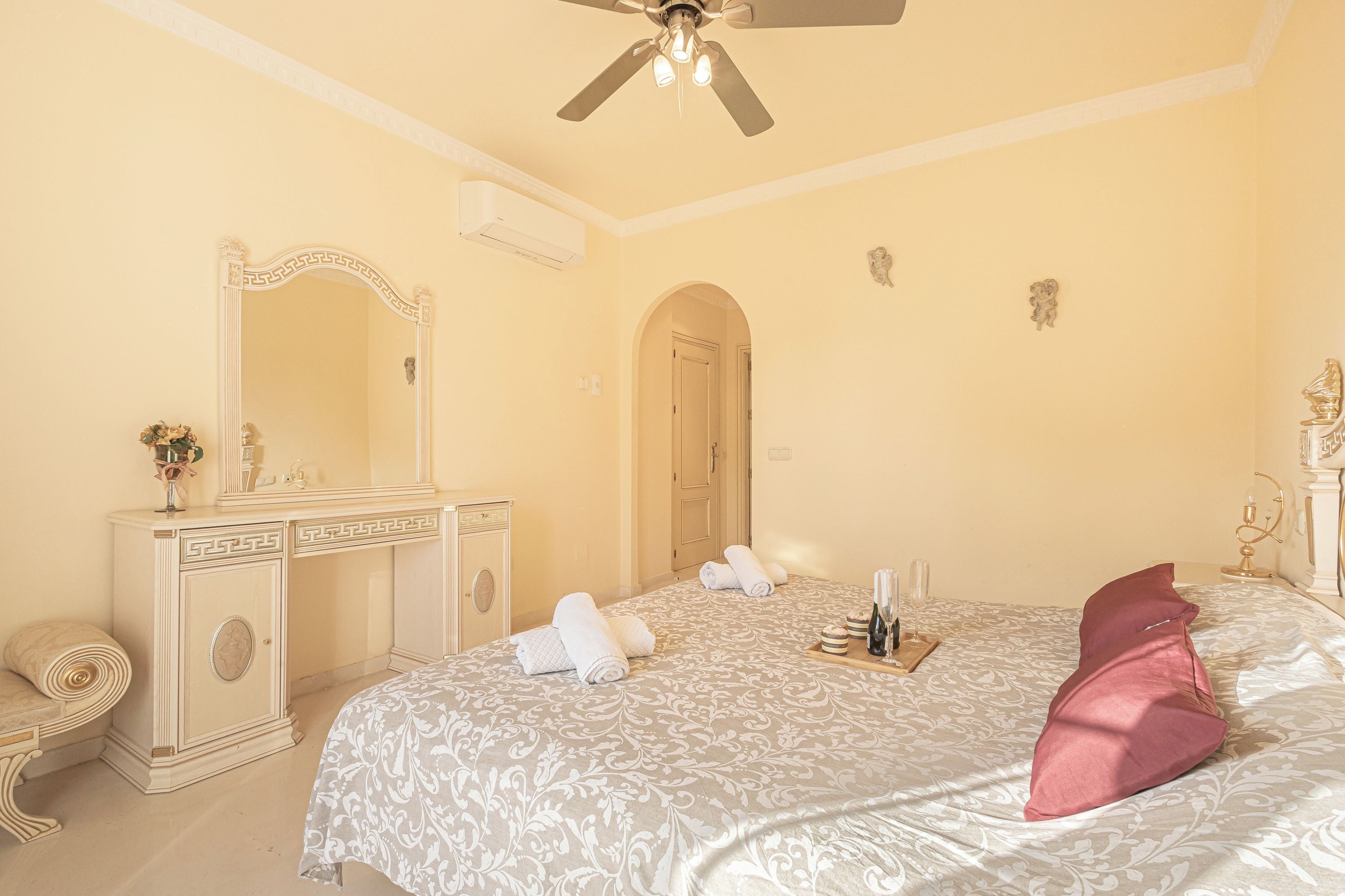 A hotel room with a bed, a vanity with a mirror, a ceiling fan, and a door leading to another room.
