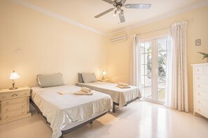 Camera da letto con due letti, una ventola a soffitto, un condizionatore d'aria, un comodino con lampada, una cassettiera e vista sull'esterno tramite una porta scorrevole in vetro.