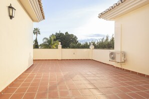 Una terrazza sul tetto con piastrelle in terracotta, un muro bianco, un condizionatore d'aria montato e vista su alberi e una palma.