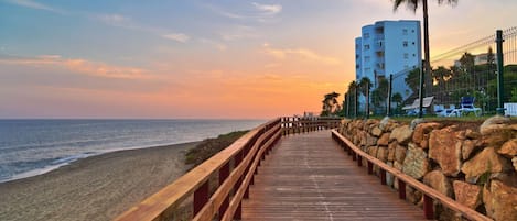 A wooden boardwalk leading to a beach, with a palm tree and a tall building in the background.