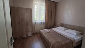 Un dormitorio con un armario de madera, una cama con ropa blanca y una ventana con cortinas traslúcidas.