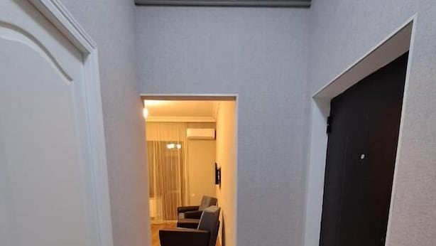 Un pasillo con una puerta oscura a la derecha, una blanca a la izquierda y una habitación al final con un sofá oscuro y cortinas.