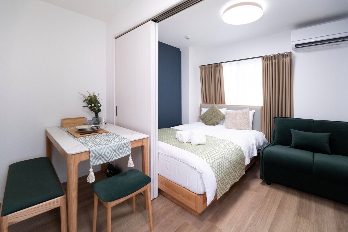 Ein Zimmer mit einem Bett, einem grünen Sofa, einem Schreibtisch mit Stuhl sowie einem kleinen Tisch mit einer Vase.
