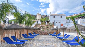 Un espacio al aire libre con suelo de piedra, sillones de descanso azules y sombrillas de paja.