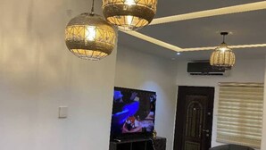 Una sala de estar con televisor, dos lámparas colgantes, un sofá y una mesa de comedor con individuales.