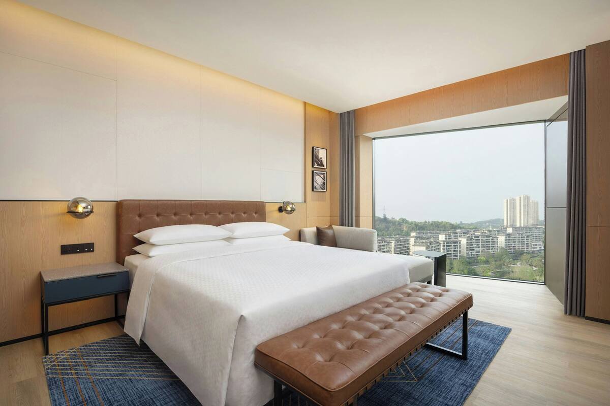 Una habitación de hotel moderna con una cama grande, un banco, una mesita de noche y vistas a la ciudad.