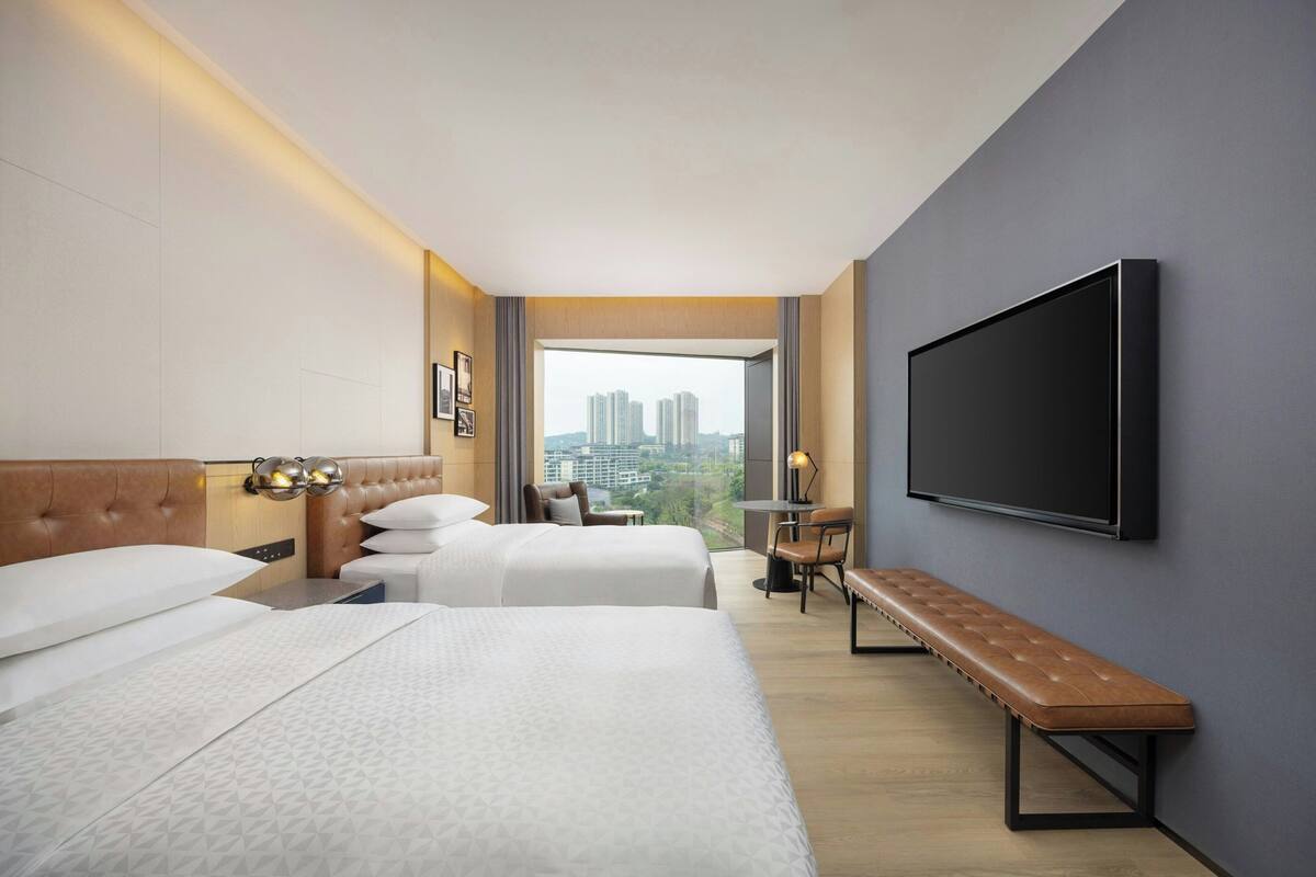 Habitación de hotel con dos camas, un banco, televisor y vista a la ciudad.