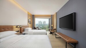 A hotel room with two beds, a bench, a TV, and a view of the city.