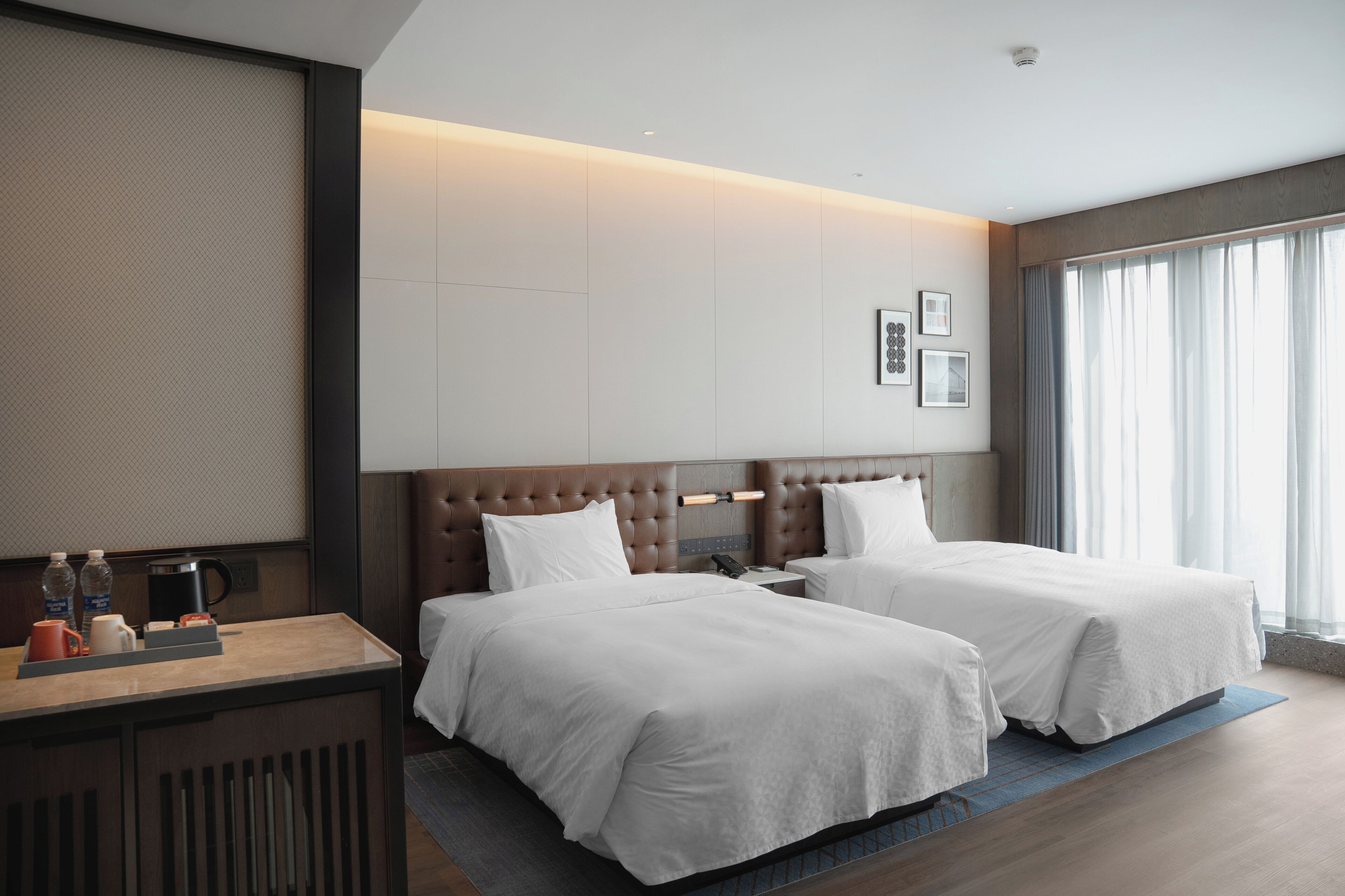 A hotel room with two beds, a desk with a coffee maker, and a large window with curtains.