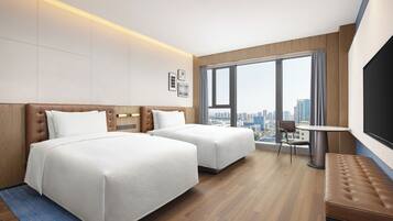 A hotel room with two beds, a small table, a bench, a TV, and a large window with a city view.