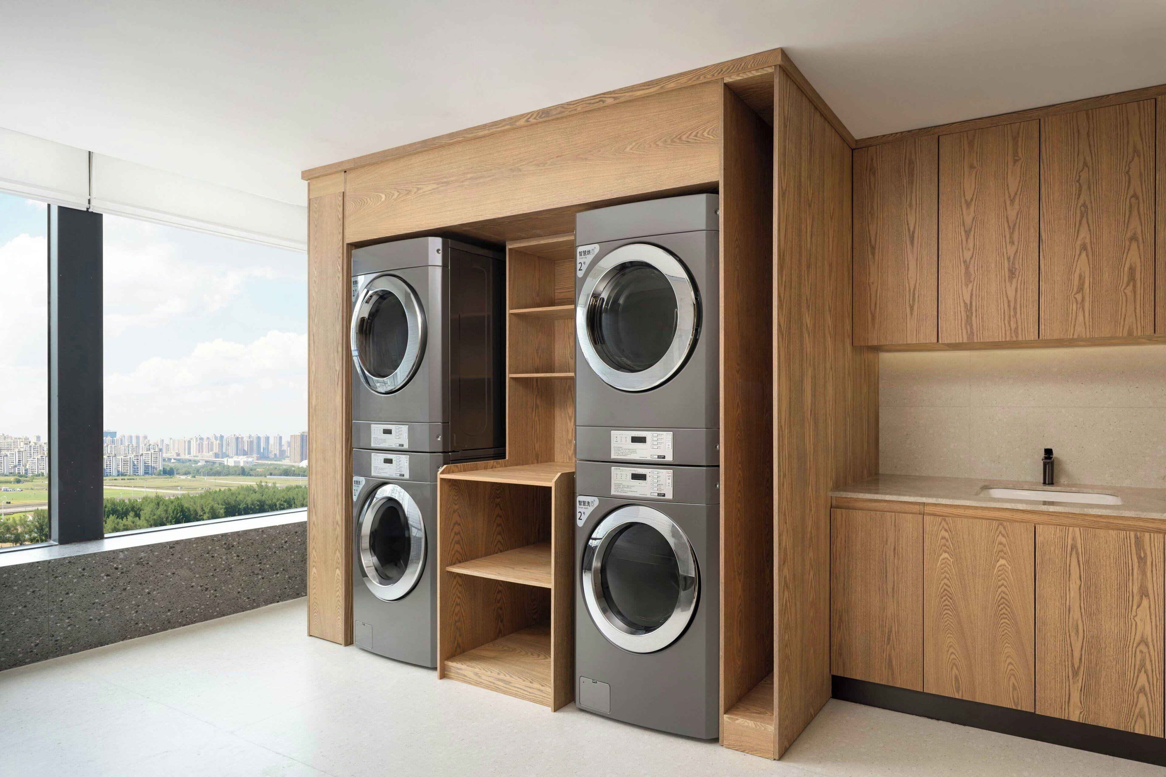 A modern laundry room with built-in washers and dryers, wooden cabinetry, and a view of a cityscape through large windows.