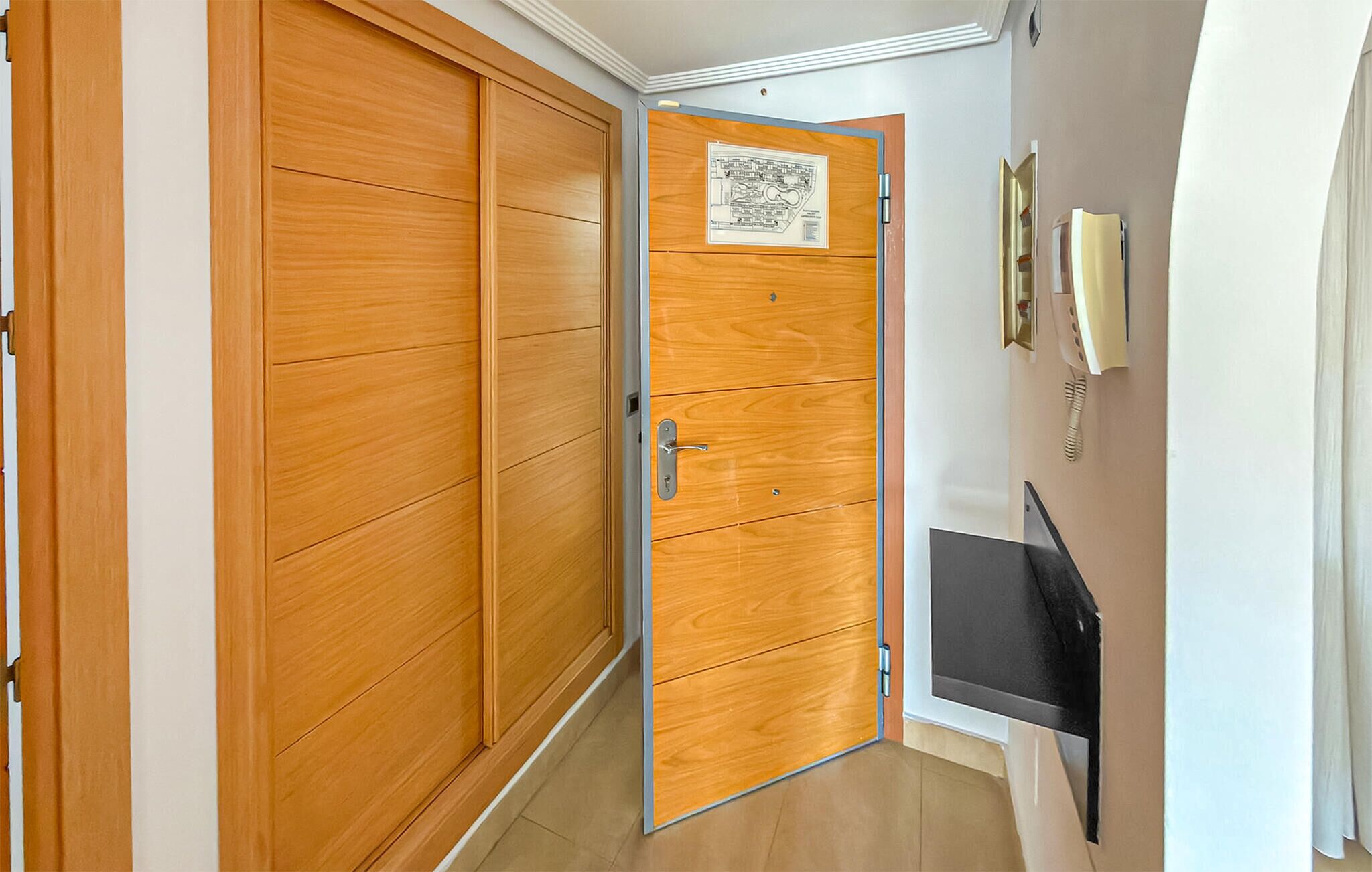 A hallway with a wooden door, a wall-mounted phone, and a small black object on the wall.