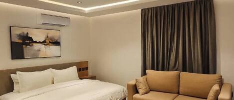 A modern hotel room with a bed, a sofa, a round coffee table, and a painting on the wall.