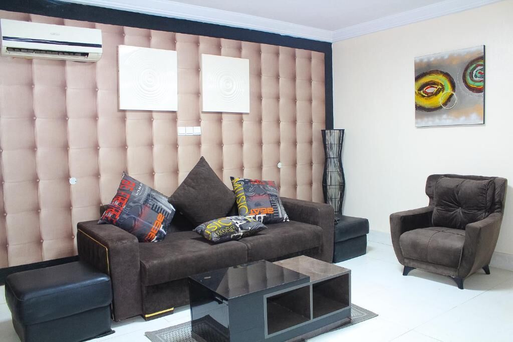 A modern living room with a tufted headboard, a brown sofa with colorful cushions, a black armchair, and a glass coffee table.