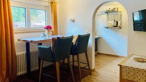 A dining area with a table, chairs, a radiator, a window with curtains, and a kitchen area with a shelf and a television. - Between blossoms & mountains, welcome to the beehive (Fischen im Allgäu)