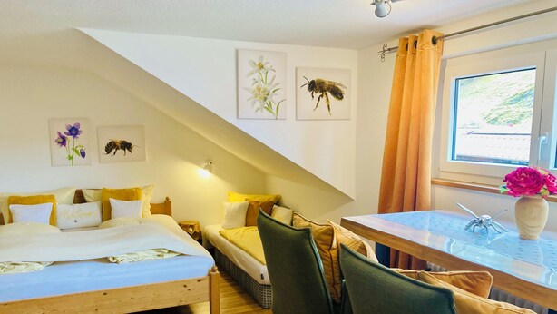 A cozy room with a bed, a small table, and a chair. There are wall art pieces and a window with curtains. - Between blossoms & mountains, welcome to the beehive (Fischen im Allgäu)