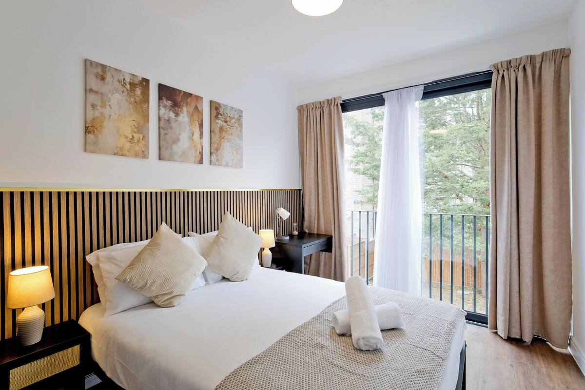 Une chambre d’hôtel avec un grand lit, une table de chevet avec une lampe, un bureau avec une lampe et une vue sur l’extérieur grâce à une porte-fenêtre coulissante.