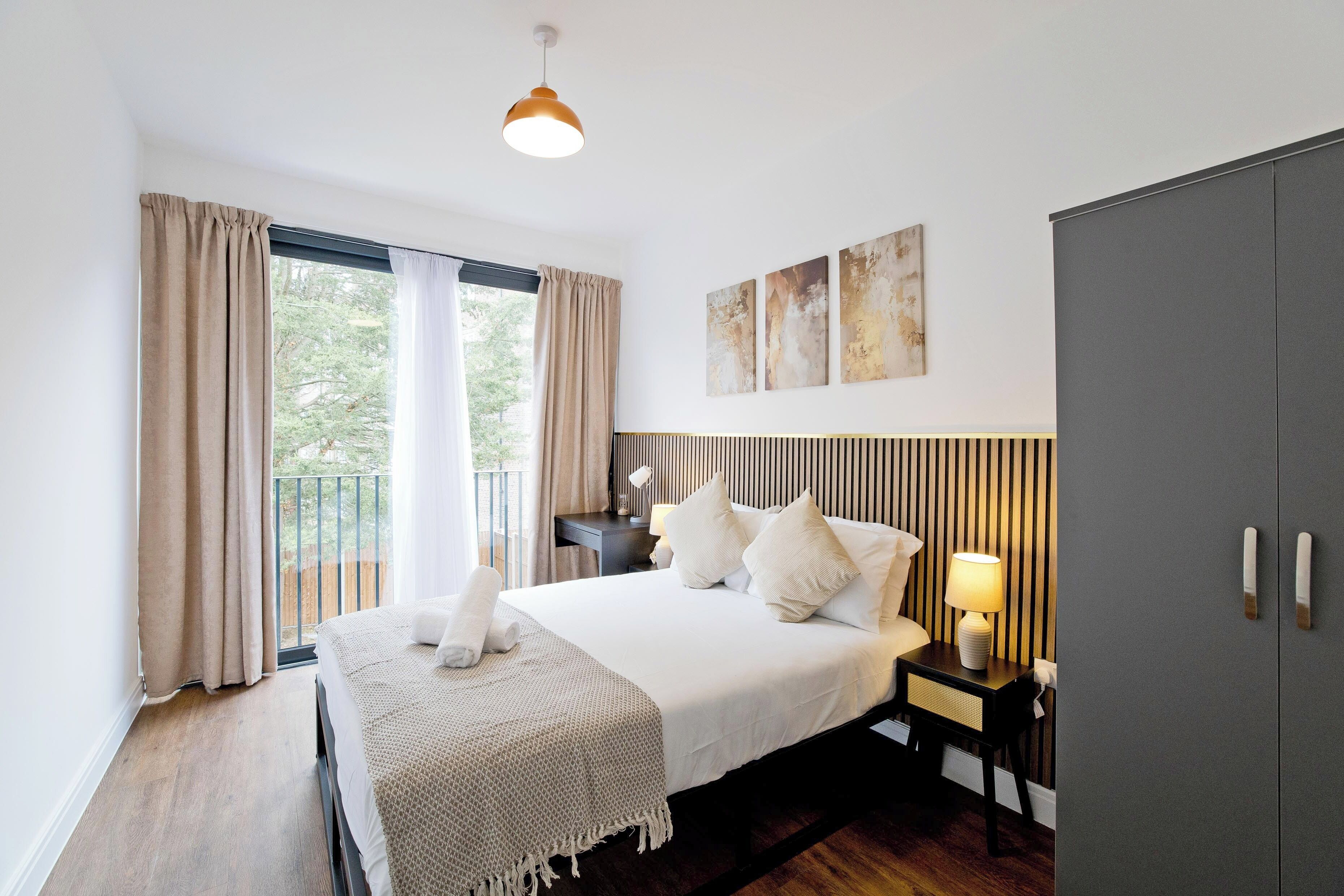 A bedroom with a bed, bedside table with a lamp, a wardrobe, and a view of greenery through the window.