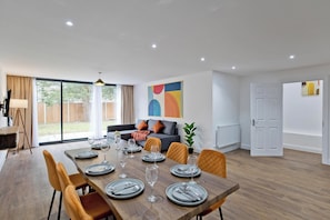 A dining area with a wooden table set for six, surrounded by orange chairs, and a modern living space with a sofa and cushions. - Imperial Woodberry Grove Apartments (London)