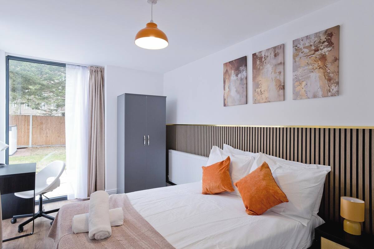 Une chambre d’hôtel moderne, équipée d’un lit, d’un bureau et d’une chaise. Les murs sont ornés de tableaux abstraits, il y a une fenêtre avec des rideaux et la chambre offre une vue sur l’extérieur.