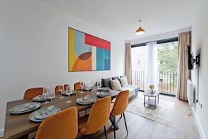 A dining area with a wooden table set for six, orange chairs, and a modern abstract painting on the wall. - Imperial Woodberry Grove Apartments (London)