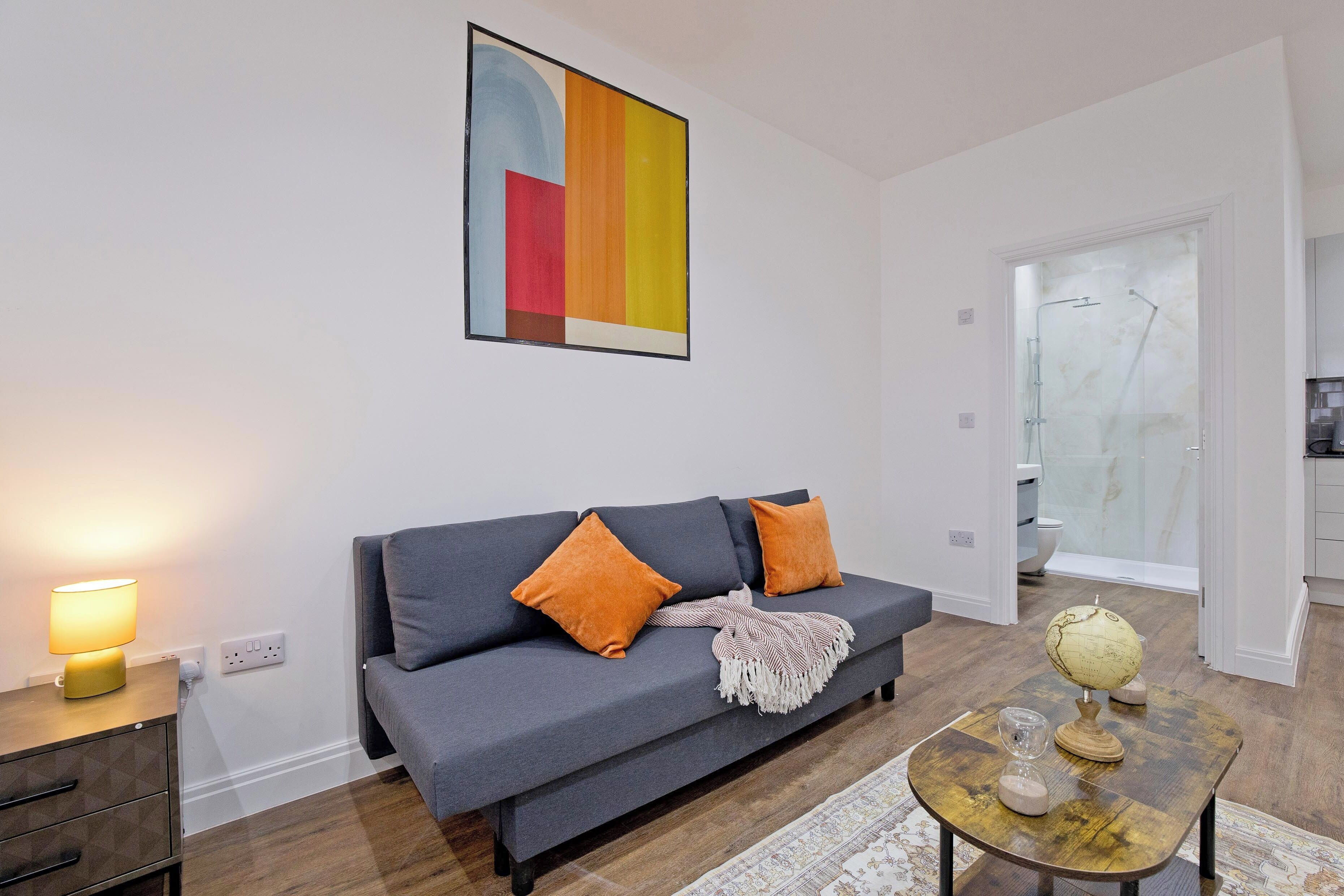 A modern living room with a grey sofa, orange cushions, a wooden side table with a lamp, and a coffee table with a globe and glasses.