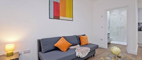 Una sala moderna con un sofá gris, cojines naranjas, una mesita de madera con una lámpara y una mesa de centro con un globo terráqueo y vasos.