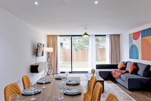 A modern living room with a dining area, large windows, and a grey sofa with cushions. - Imperial Woodberry Grove Apartments (London)