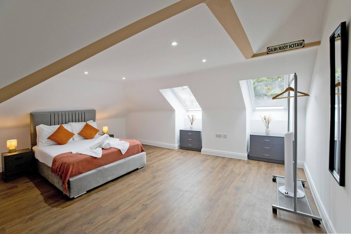 Une chambre avec un plafond mansardé, un lit avec des coussins orange, une table de chevet avec une lampe, une commode et un miroir.