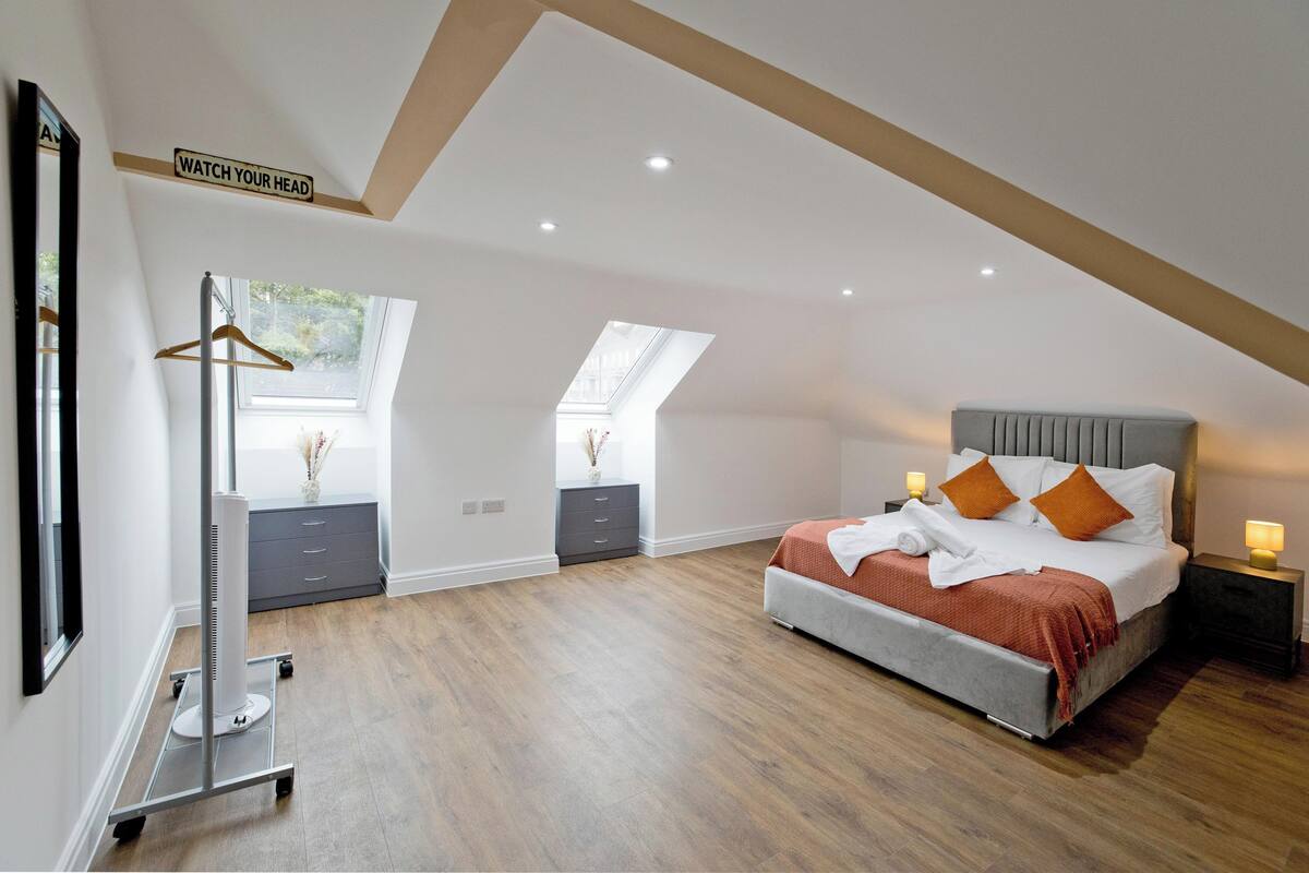 Une chambre avec un plafond en pente, un lit avec des coussins orange, une table de chevet avec une lampe et un panneau décoratif fixé au mur.