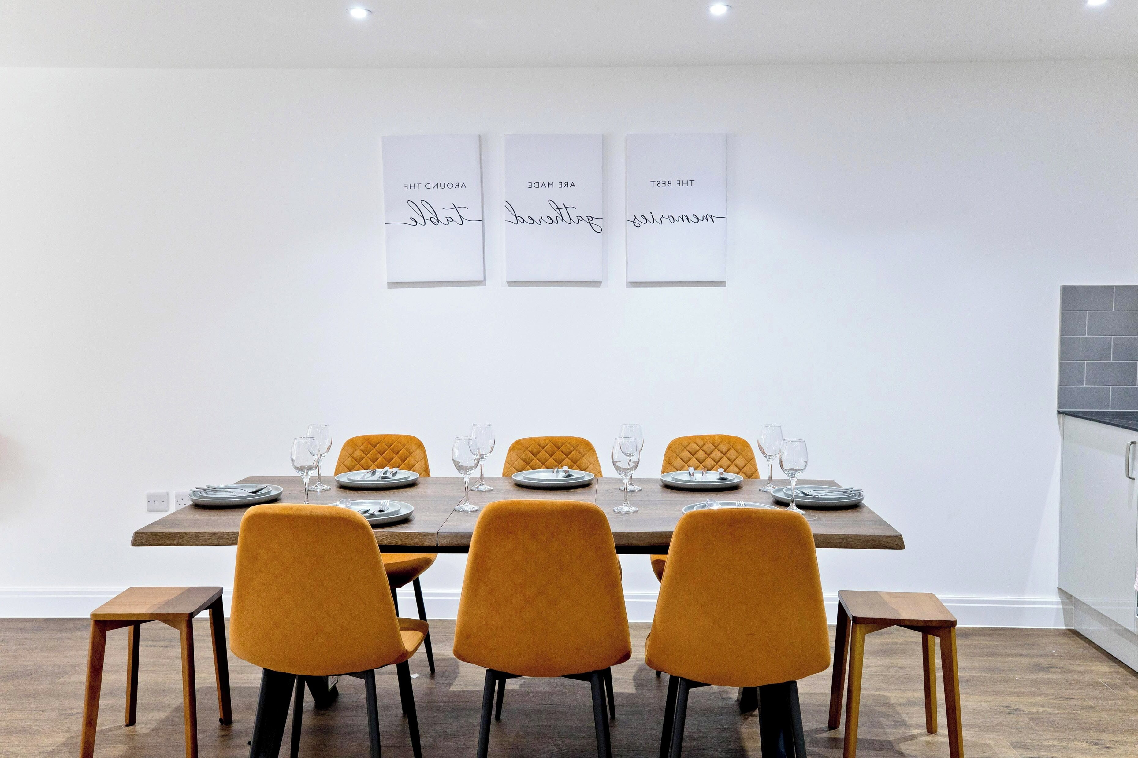 Un coin repas avec une table en bois dressée pour six personnes, entourée de six chaises de couleur moutarde. Trois panneaux encadrés avec du texte sont accrochés au mur au-dessus de la table.