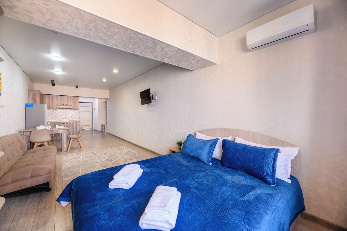 A modern hotel room with a large bed, a sofa, a desk, and a kitchenette.