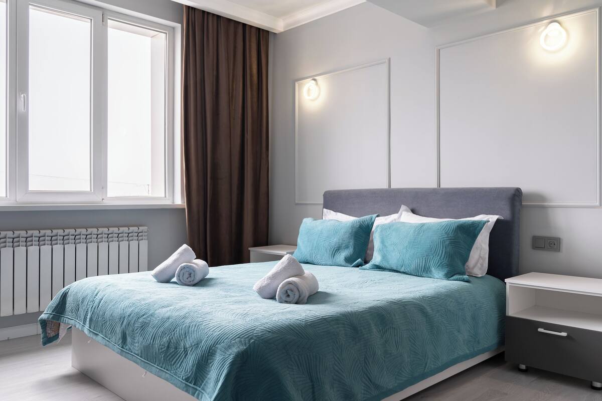 A modern bedroom with a large bed, two bedside tables, a radiator, and a window with curtains.