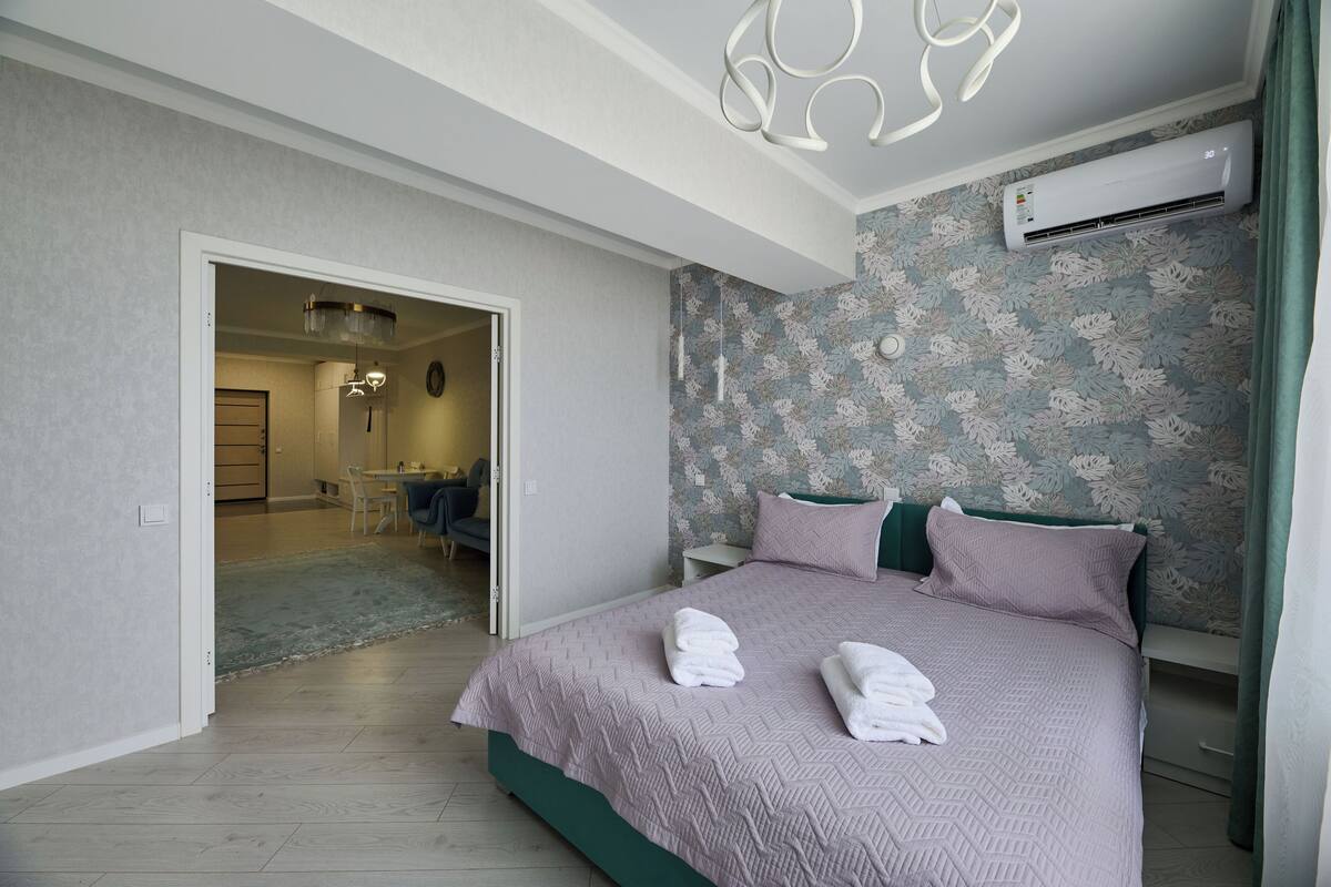 A bedroom with a bed, a nightstand, an air conditioner, and a bathroom visible through an open door.