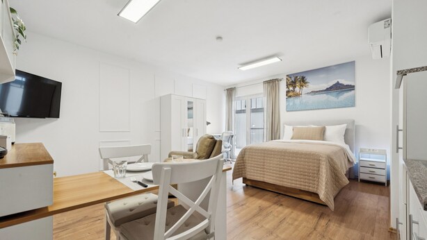 Une chambre d’hôtel moderne avec un plancher en bois, un lit recouvert d’une couette beige, un bureau avec une chaise, un téléviseur fixé au mur et une fenêtre avec des rideaux.