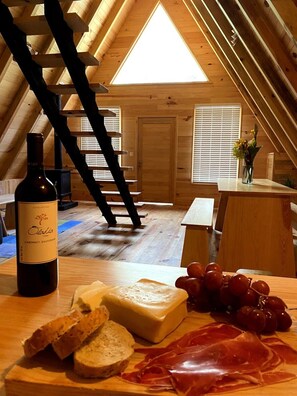 A wooden interior with a sloped ceiling, a bottle of wine, cheese, bread, grapes, and cured meat on a table.