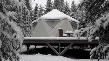 Uma tenda coberta de neve com uma mesa e cadeiras, rodeada por árvores carregadas de neve.