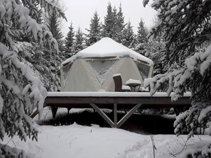 A snow-covered tent with a table and chairs, surrounded by snow-laden trees.