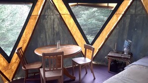 A room with a wooden table and chairs, a small bedside table with a lamp, and a bed.