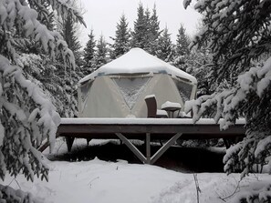 A snow-covered tent with a table and chairs, surrounded by snow-laden trees.